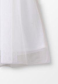 Witte tule stof met een zachte, doorschijnende textuur; gelaagd met een delicate, luchtige val aan de zoom, wat een lichte uitstraling creëert.