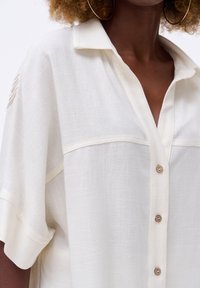 Witte, linnenmix blouse met een kraag, korte mouwen en een knoopsluiting aan de voorkant. Kenmerkend zijn de textuur van de stof en de subtiele naaifittingen.