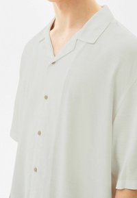 Camisa de manga corta gris claro, corte relajado, con cuello con solapa y cinco botones beige en la parte delantera, textura de tela lisa.