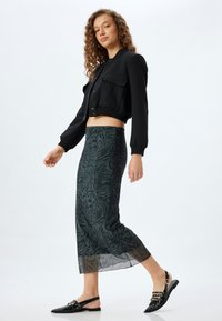 Zwarte cropped jack met knoopsluiting, gecombineerd met een doorschijnende groene rok met een tijgerstrepenpatroon, en zwarte slingback schoenen.