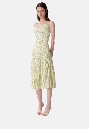 Femme portant une robe midi jaune à fleurs avec bordure en dentelle et bretelles nouées aux épaules, associée à des talons ouverts blancs.