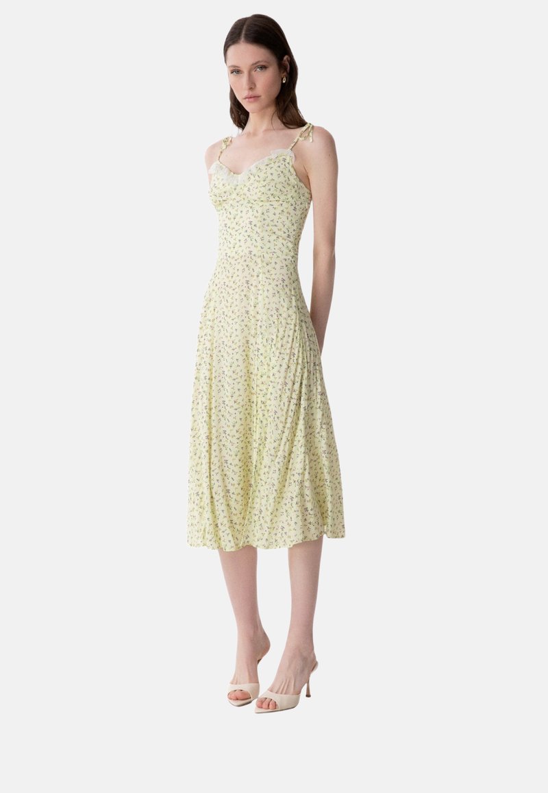 Frau trägt ein gelbes Midikleid mit Blumenmuster, Spitzenbesatz und gebundenen Trägern an den Schultern, kombiniert mit weißen offenen High Heels.