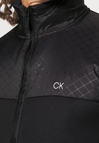 Czarna kurtka na zamek błyskawiczny z subtelnym wzorem w diamenty na górze i białym logo "CK" na lewym klatce piersiowej, noszona przez osobę z kręconymi włosami.