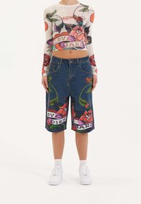 Pantalones cortos de mezclilla con bordados florales, que presentan colores llamativos y texto gráfico. Combinados con una blusa de manga larga transparente adornada con un estampado floral.