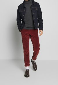 Dunkle Jeansjacke, grauer Rollkragenpullover, rote Cordhosen und braune Schuhe mit weißen Socken. Accessoires minimalistisch mit glatten Texturen.