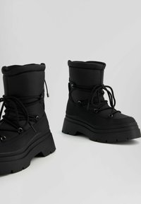 Svarta vadderade ankelstövlar med rund tå, tjocka gummisulor och metallöglor för snörning. Har strukturerad tyg och framträdande sömmar.