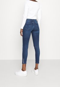 Smala blå denimjeans med mid rise midja, bakfickor och subtila sömdetaljer; matchade med vita sneakers och en vit långärmad topp.