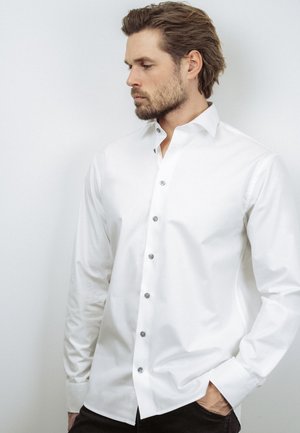 Brodaty mężczyzna o jasnobrązowych włosach, ubrany w białą koszulę na guziki, spoglądający na bok, z jedną ręką w kieszeni, na białym tle.