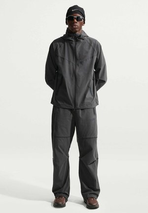 Mężczyzna ubrany w ciemnoszary dres Nike, czarną opaskę na głowę Nike, okulary przeciwsłoneczne i brązowe buty trekkingowe, stojący na jednolitym jasnym tle.