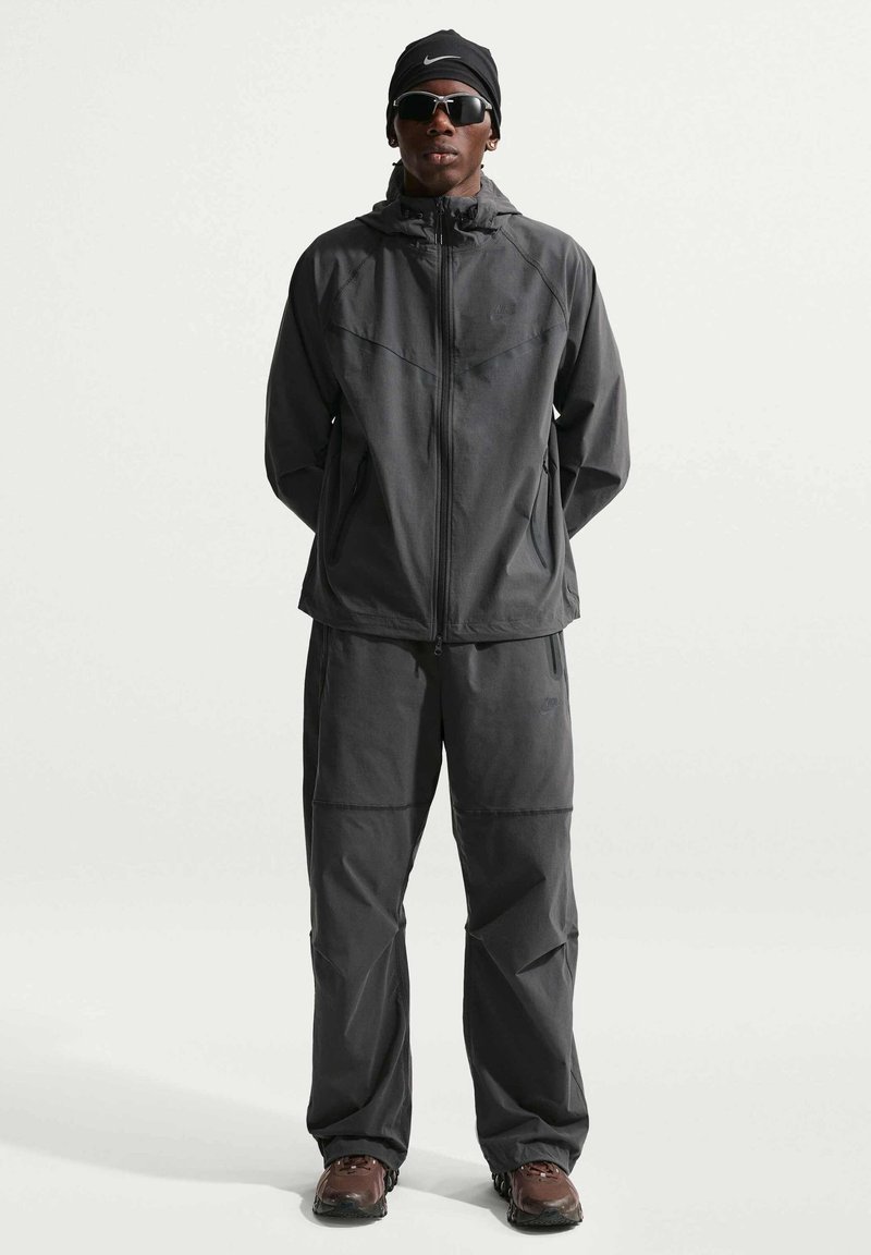 Man wearing dark gray Nike tracksuit, black Nike headband, sunglasses, and brown hiking shoes, standing against plain light background.