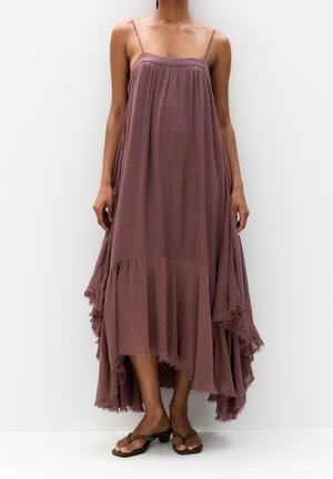 Robe longue et fluide en mauve atténué, avec des fines bretelles, un corsage froncé et un ourlet effiloché. Portée avec des tongs simples.