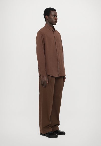 Chemise marron à manches longues avec un design à boutons, poche poitrine, associée à un pantalon marron ample. Des chaussures noires complètent l'ensemble.