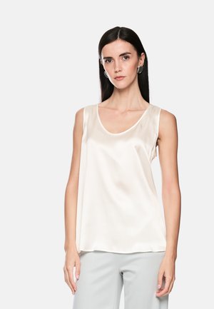 Woman with long dark hair wearing silver hoop earrings, a sleeveless white satin top, and light gray pants against a plain background.