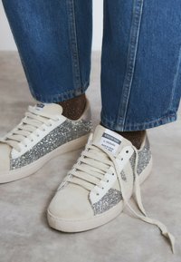 Zapatillas blancas con detalles de purpurina plateada en los lados y detalles en ante color crema. Los cordones son planos y la suela de goma.