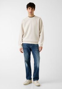 Sweat-shirt couleur crème avec un col arrondi et des poignets côtelés, associé à un jean bleu clair délavé et des baskets blanches.