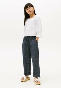 Blusa bianca a maniche lunghe con dettagli testurizzati, abbinata a pantaloni a gamba larga a strisce blu navy e sandali bianchi con platform. La modella sta in piedi con le mani in tasca.