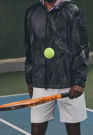 Person iført en mørk mønstret jakke og hvide shorts holder et orange tennisbat, mens en gul tennisbold hopper foran.