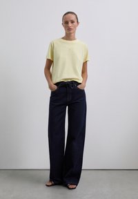 Žluté bavlněné tričko s kulatým výstřihem, spárované s vysokými širokými džínami z tmavé rifloviny. Sandály s jednoduchým designem pásku.