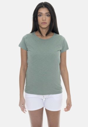 Donna con lunghi capelli scuri che indossa una maglietta verde a maniche corte e pantaloncini bianchi, in piedi dritta su uno sfondo bianco semplice.