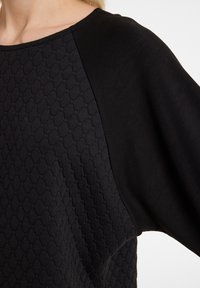 Schwarzer Pullover mit einem strukturierten, hexagonalen Muster auf einer Seite und glattem Stoff auf der anderen Seite, mit rundem Halsausschnitt und lockerer Passform.