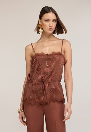 Femme aux cheveux courts bruns portant un caraco en satin couleur rouille orné de motifs de feuilles et un pantalon assorti, accessoirisée avec des boucles d'oreilles et des bracelets en or.