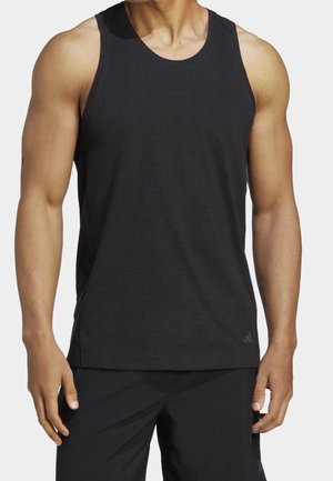Muskelöser Mensch trägt ärmelloses schwarzes sportliches Tanktop und schwarze Shorts und steht mit entspannten Armen an den Seiten.