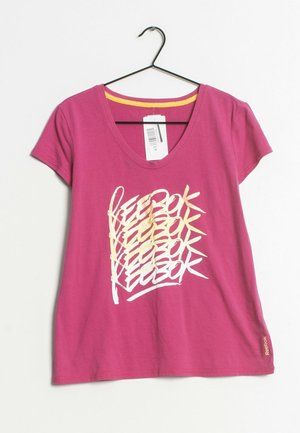 Różowy T-shirt z krótkim rękawem z powtarzającym się kremowo-żółtym wzorem tekstu "Reebok", żółtym wewnętrznym wykończeniem kołnierza i białą metką cenową zawieszoną na czarnym wieszaku.