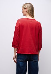 Felpa rossa con maniche a tre quarti, dettagli a coste sulle maniche e vestibilità rilassata, abbinata a jeans di denim blu scuro.