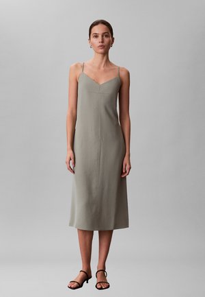 Femme debout portant une robe midi grise sans manches avec de fines bretelles et un décolleté en V, chaussée de sandales noires à talons avec des lanières, sur un fond uni.