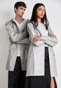 Deux mannequins portent des vestes imperméables gris clair, dotées de boutons-pression et de poches à l'avant. Les vestes ont une coupe décontractée et des capuches avec cordon de serrage.