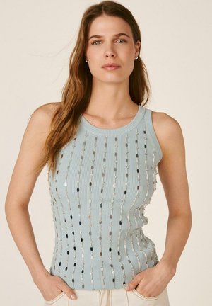 Mujer con cabello largo y castaño que lleva una blusa sin mangas azul claro con rayas verticales de lentejuelas plateadas, de pie con las manos en los bolsillos del pantalón.