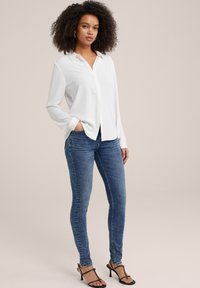 Camisa blanca de manga larga y botones combinada con jeans ajustados azules y tacones negros con tiras. Diseño simple, tejido ligero, corte clásico.