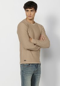 Beige hosszúujjú póló bordás textúrával, kerek nyakkivágással és egy kis logócímkével az alján. Kék farmerral párosítva.