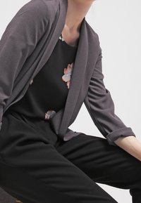 Šedý, strukturovaný cardigan přeložený přes černý květinový top, s volným střihem a ohrnutými rukávy, s černými kalhotami níže.