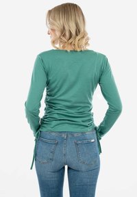Top teal a maniche lunghe con lati arricciati e lacci, abbinato a jeans in denim di lavaggio medio con un classico design a cinque tasche.
