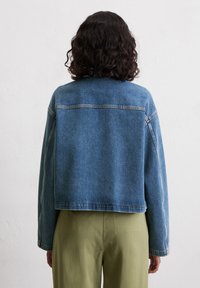 Osoba z kręconymi włosami widoczna z tyłu, ubrana w skróconą dżinsową kurtkę na oliwkowo-zielone spodnie, na tle jasnego tła.