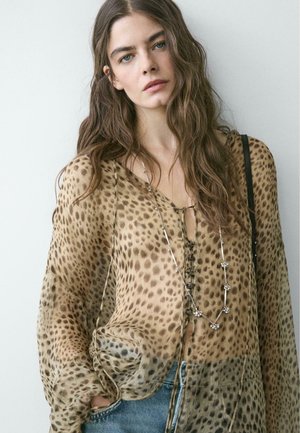 Donna con capelli lunghi e ondulati che indossa una camicetta trasparente con stampa leopardata, una collana d'argento e jeans blu, in piedi davanti a uno sfondo chiaro e uniforme.