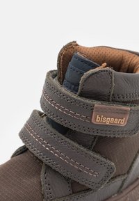 Botte montante en cuir gris avec des accents en textile marron, deux straps Velcro réglables et un patch de marque sur le côté. Détails de la surface texturée.