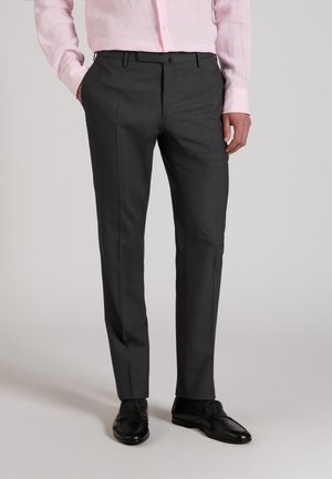 Mężczyzna w ciemnoszarych dopasowanych spodniach, czarnych eleganckich butach i jasnoróżowej koszuli na guziki, z rękami w kieszeniach.