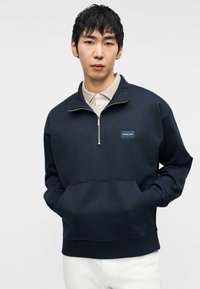 Σκούρα ναυτικό μπλουζάκι με φερμουάρ στη μέση, που διαθέτει εμπρόσθια τσέπη, ριμπ μανσέτες και μια μικρή ετικέτα με το λογότυπο στο στήθος.