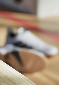 Weiße Sportschuhe mit schwarzen Akzenten auf einer Holzoberfläche, teilweise verschwommen, wobei die strukturierte Sohle und das schlichte Design zu sehen sind.