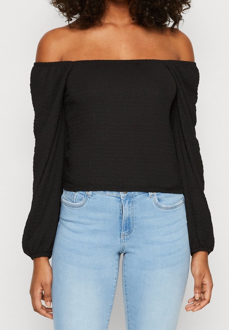 Musta off-the-shoulder -paita, jossa on puffatut pitkät hihat, teksturoitu kangas ja tiukka vartalonmyötäinen malli, yhdistettynä vaaleansinihin farkkuihin.