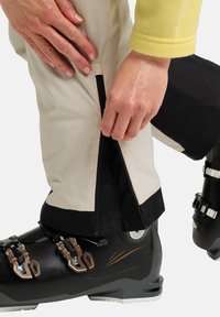 Sneeuwbroeken van beige stof met een zwarte boord, een zijritsdetail en een close-up van een skiboot met metalen hardware en accenten.