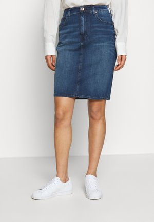 Person iført en knælængde blå denimnederdel, hvid langærmet skjorte og hvide sneakers, stående mod en ensfarvet lys baggrund.