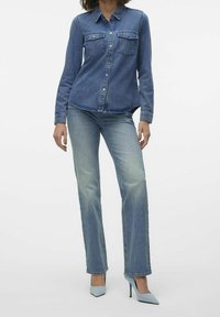 Nainen, joka pukeutuu siniseen farkkupaitaan, vaaleansinisiin farkkuihin ja vaaleansinisiin korkeakorkoisiin kenkiin, seisoo yksinkertaista valkoista taustaa vasten.