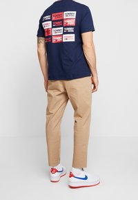 Navy blue t-shirt with multiple red and white "TOMMY JEANS" logos on the back, paired with beige pants and white sneakers with blue accents.