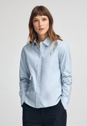Camisa de botones azul clara, hecha de una tela suave, con cuello, mangas largas y un pequeño logo en el lado izquierdo del pecho.