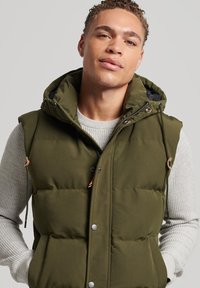 Gepolsterte olivgrüne Weste mit Kapuze, ausgestattet mit Druckknöpfen und Kordelzügen, die über einem hellgrauen strukturierten Pullover getragen wird.