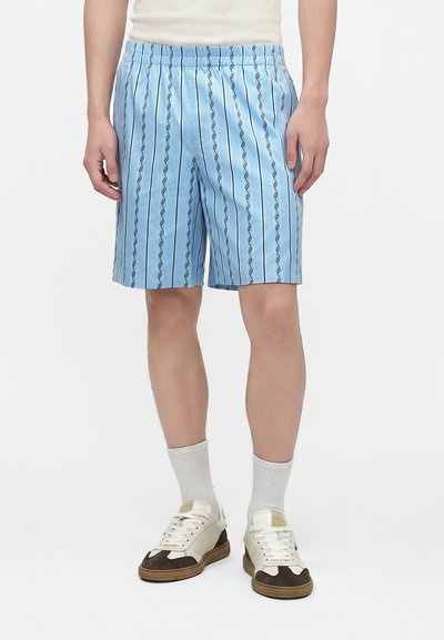 Un homme porte un short rayé bleu clair, des chaussettes blanches et des baskets blanches avec des touches marron, debout devant un fond blanc uni.