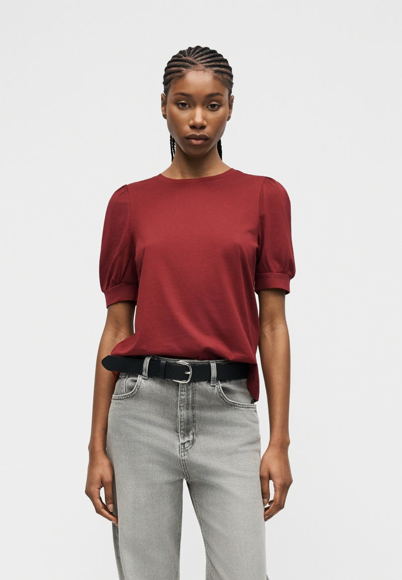 Γυναίκα με πλεγμένα μαλλιά που φοράει κόκκινο μπλουζάκι με κοντά μανίκια, μπηγμένο σε ψηλόμεσα ανοιχτογκρι τζιν με μαύρη ζώνη, στέκεται μπροστά σε γκρι φόντο.